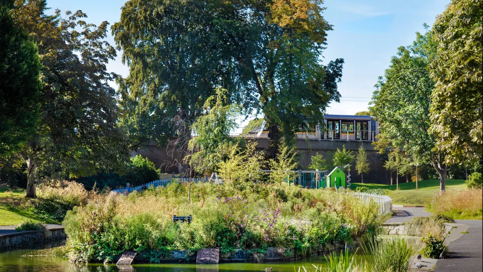 A lush urban park with a pond in the foreground surrounded by tall grasses and flowering plants. Behind the pond, there is a small green playhouse and a fenced playground area. Mature trees with dense green foliage frame the scene, and a modern tram passes on an elevated track in the background under a clear blue sky.