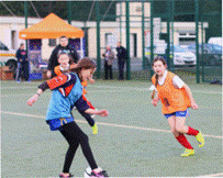 A group of young players actively participating in a football match on an outdoor artificial turf pitch. Two players in orange bibs are moving towards the ball while another player in a blue bib is dribbling. A coach or spectator stands near an orange canopy in the background, with fencing and buildings visible behind the pitch.