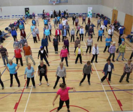 A large group of people participating in a fitness or exercise class inside a sports hall. The participants are spaced out across a wooden gym floor marked with red and white court lines, following the instructor at the front. Chairs are arranged along the walls, and a basketball hoop is visible in the background.