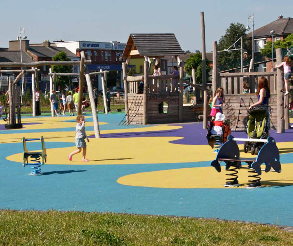 A colourful outdoor playground with bright blue and yellow rubber flooring. The play area features wooden climbing structures, slides, and rope bridges, with spring-mounted dolphin-shaped seats in the foreground. Several children are playing while adults supervise, including one person pushing a stroller. Houses and a commercial building are visible in the background under a clear blue sky.