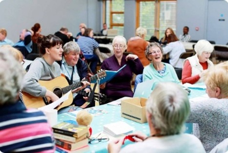 A community gathering in a bright indoor space with several tables and chairs. People are seated in groups, engaging in conversation and activities. One person is playing an acoustic guitar while others listen and interact. The tables have books, papers, and small items, suggesting a social or creative event. Large windows in the background let in natural light