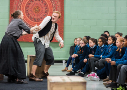 A live theatre performance taking place in a school or community hall. Two actors in period-style costumes are performing in front of an audience seated in rows. The audience consists of students in uniform watching attentively. The background features a green wall and a decorative tapestry hanging behind the performers