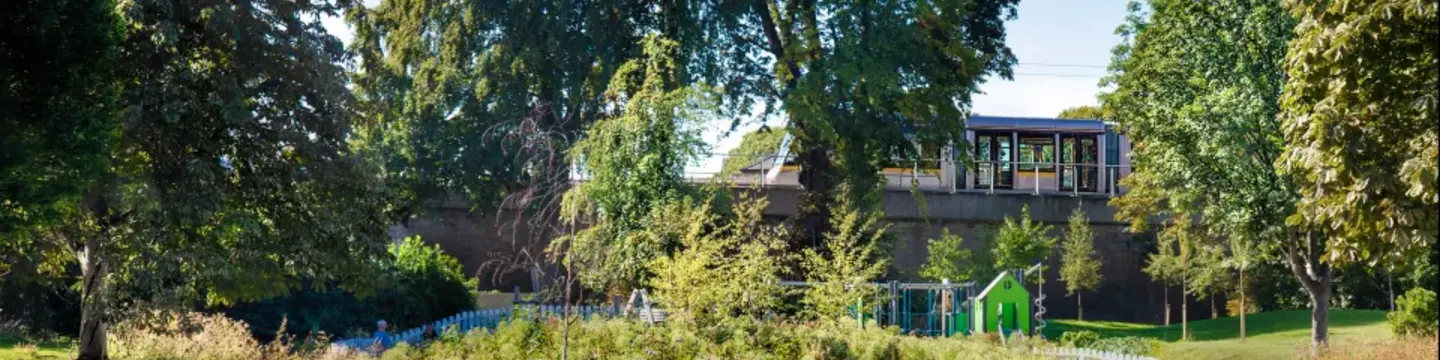 A lush urban park with a pond in the foreground surrounded by tall grasses and flowering plants. Behind the pond, there is a small green playhouse and a fenced playground area. Mature trees with dense green foliage frame the scene, and a modern tram passes on an elevated track in the background under a clear blue sky.