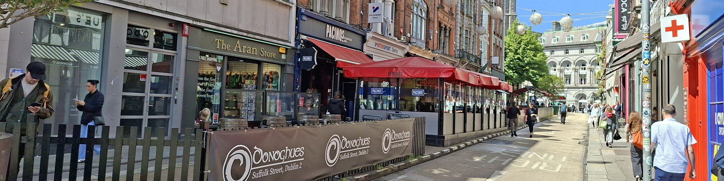Photo of Suffolk Street with restaurants, outdoor seating, hanging lights and pedestrians, framed by historic red-brick buildings and a church spire in the background.