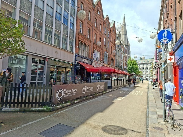 Photo of Suffolk Street with restaurants, outdoor seating, hanging lights and pedestrians, framed by historic red-brick buildings and a church spire in the background.